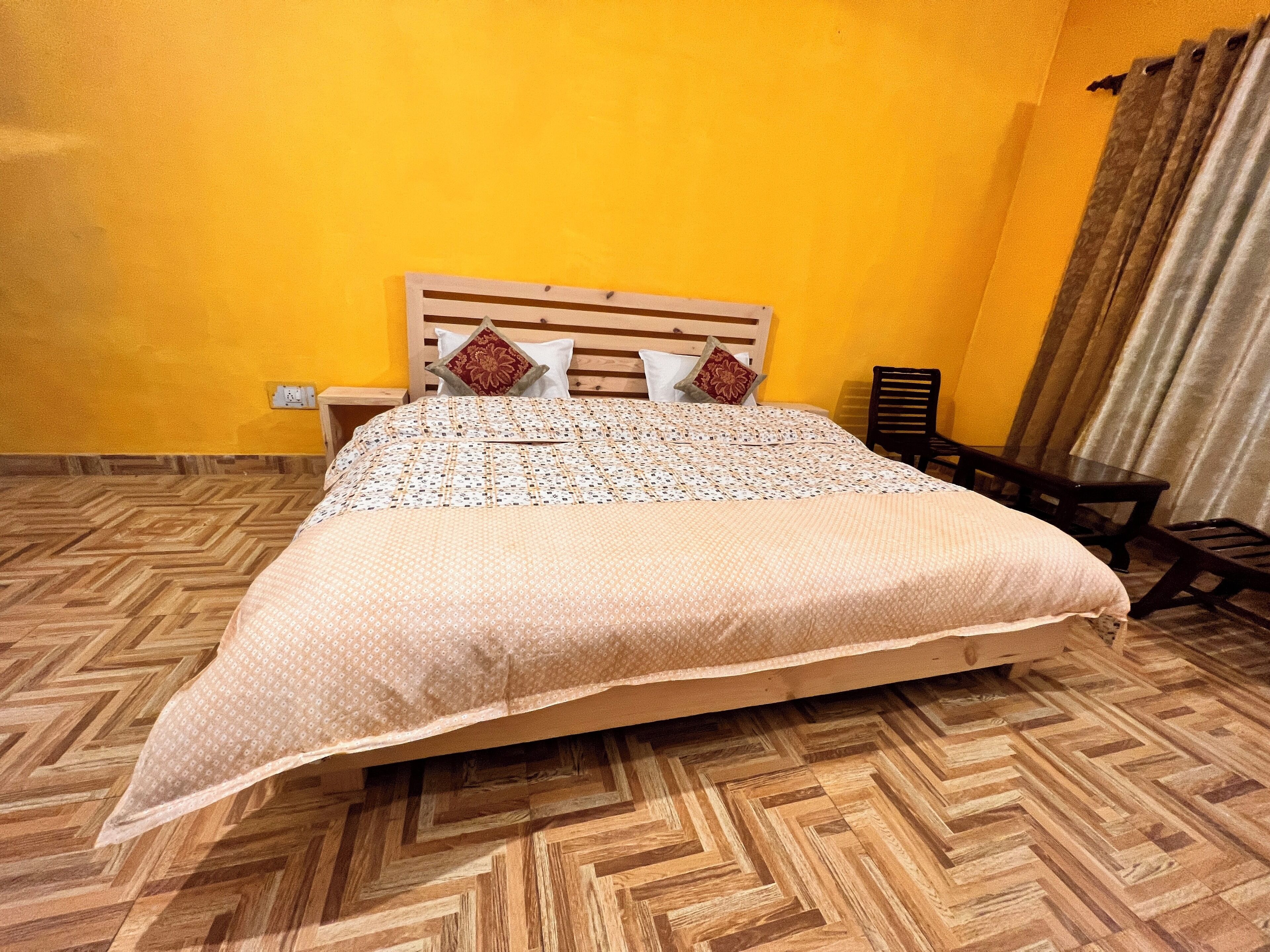 Un letto con copriletto decorato, una testata in legno e due cuscini. Si nota un comodino con una lampada, una sedia di legno e una finestra con tende.