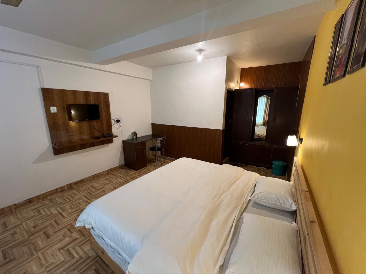 A hotel room with a bed, a desk, a chair, a TV mounted on the wall, and a wooden headboard.