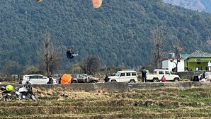 A mountainous landscape with a paraglider in the air, a group of vehicles, and a few people on the ground. - Uday Homestay (Baijnath)