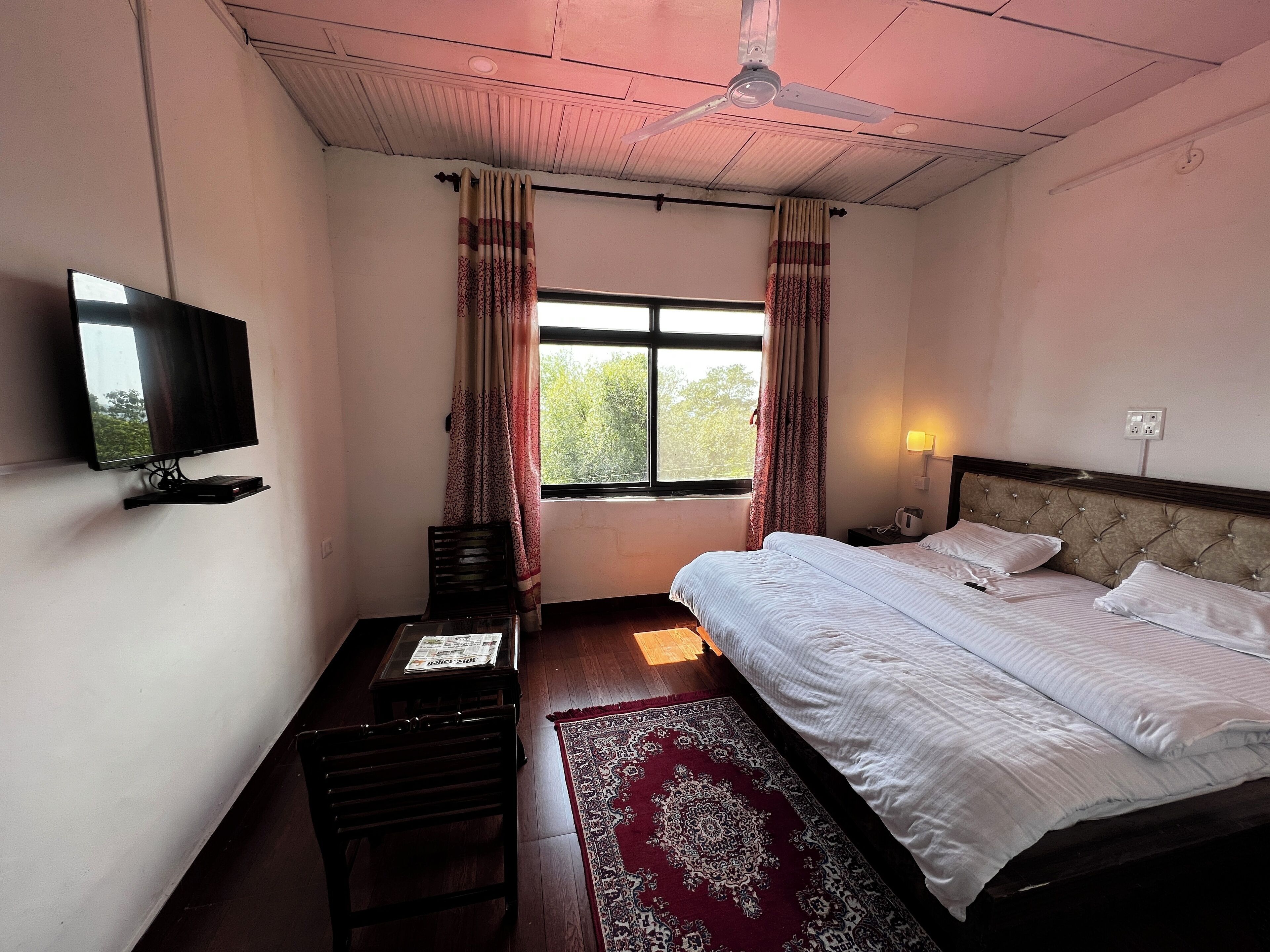 A bedroom with a bed, a television on a stand, a window with curtains, a ceiling fan, and a nightstand with a lamp.