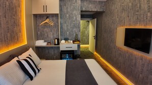 Habitación de hotel con cama, televisor, minibar, zona de estar con mesa y sillas, y un pasillo que lleva a un baño.