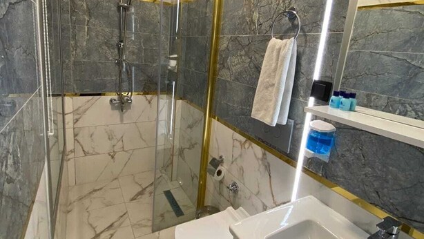 Un baño con ducha de vidrio, inodoro blanco, lavamanos blanco y espejo con marco dorado.