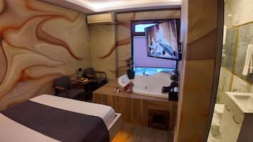 Une chambre d’hôtel comprenant un lit, un bureau, une chaise, une grande fenêtre, une télévision, une baignoire et une salle de bain avec des toilettes et un lavabo.
