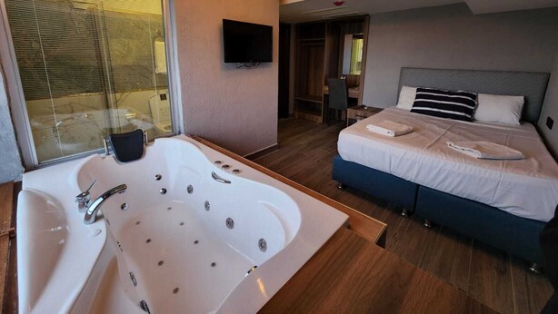 A modern hotel room with a large bathtub, a bed with two pillows, a flat-screen TV, and a walk-in closet. - Martin City Hotel (Bursa)