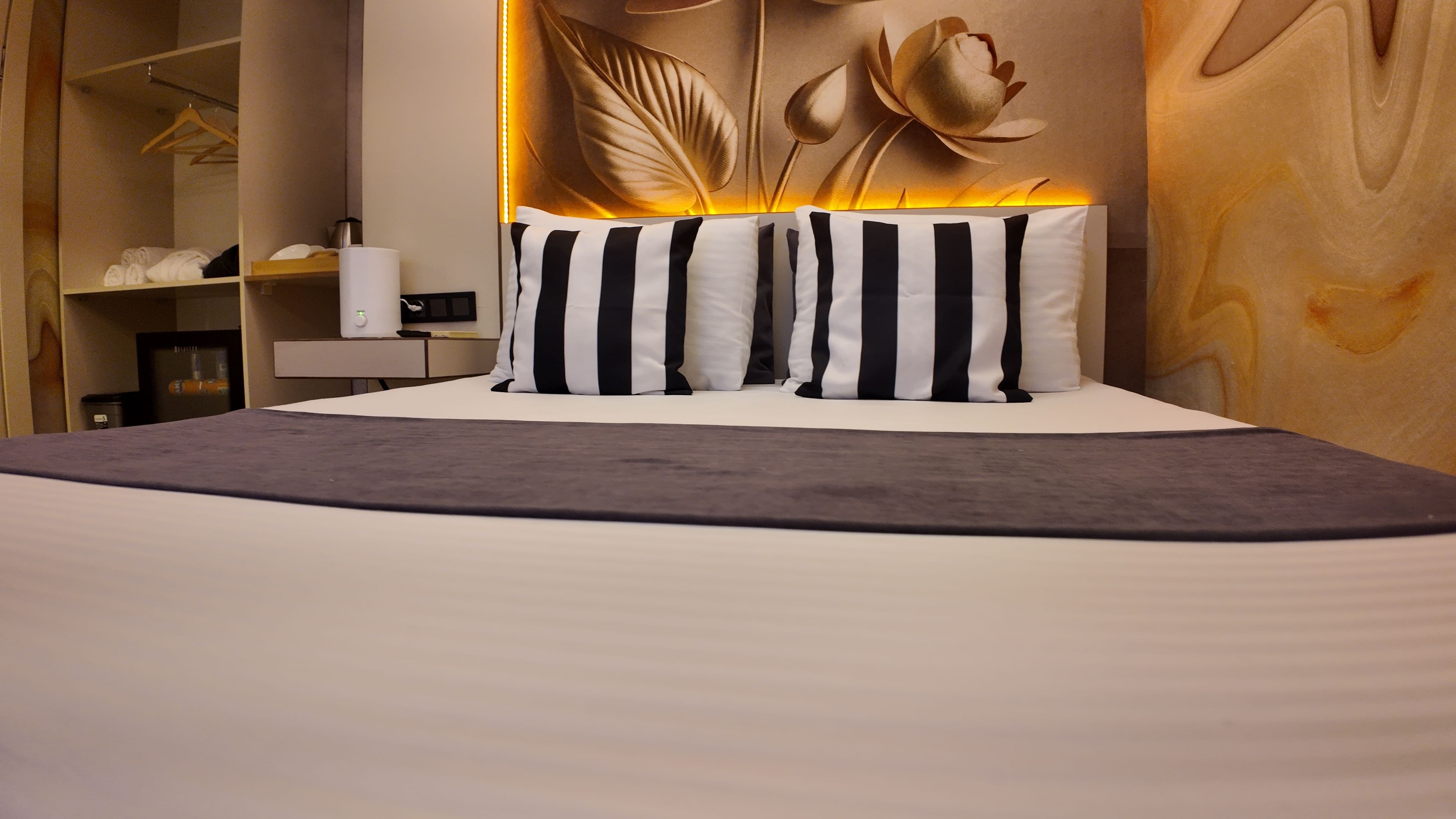 A hotel room with a bed, pillows, a bedside table, a lamp, a wardrobe, and a wall with a floral design.