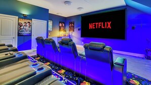 A home theater room with a large screen displaying "NETFLIX," multiple reclining seats, and posters on the walls.