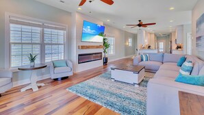 A spacious living room with hardwood floors, a large flat-screen TV, ceiling fans, and a cozy seating area with a sofa and armchairs.