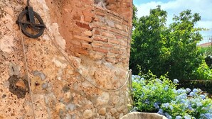 Un pozo de piedra con una cadena metálica, un muro de ladrillo y una maceta con flores rojas.