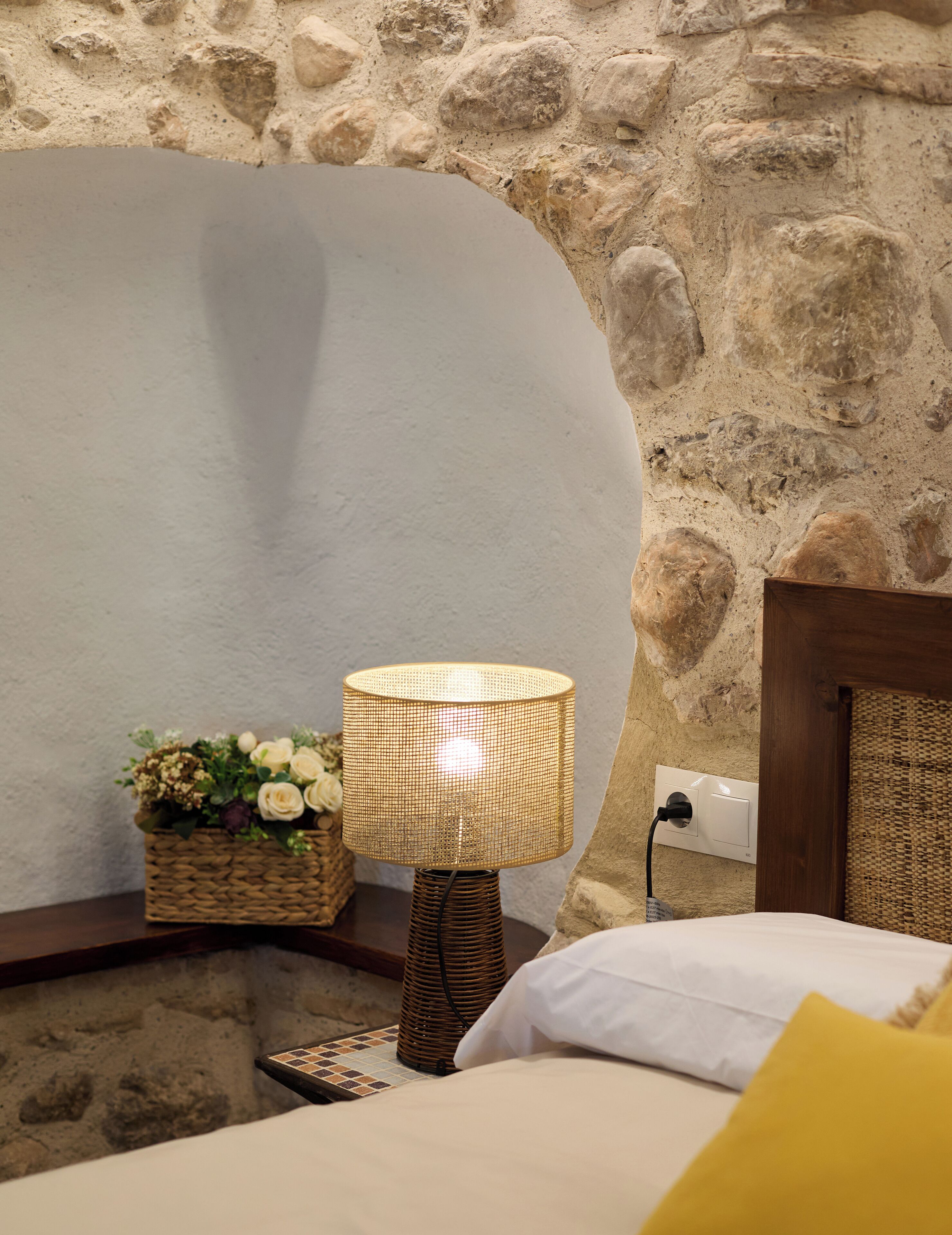 Un lit avec des oreillers blancs et jaunes, une lampe en osier et un panier de fleurs posé sur un mur en pierre.