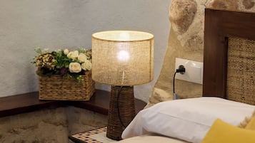 Un lit avec des oreillers blancs et jaunes, une lampe en osier et un panier de fleurs posé sur un mur en pierre.