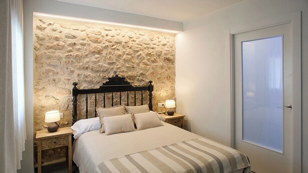 Un dormitorio con pared de piedra, cama con colcha a rayas, dos lámparas de noche y una puerta con panel de vidrio esmerilado.