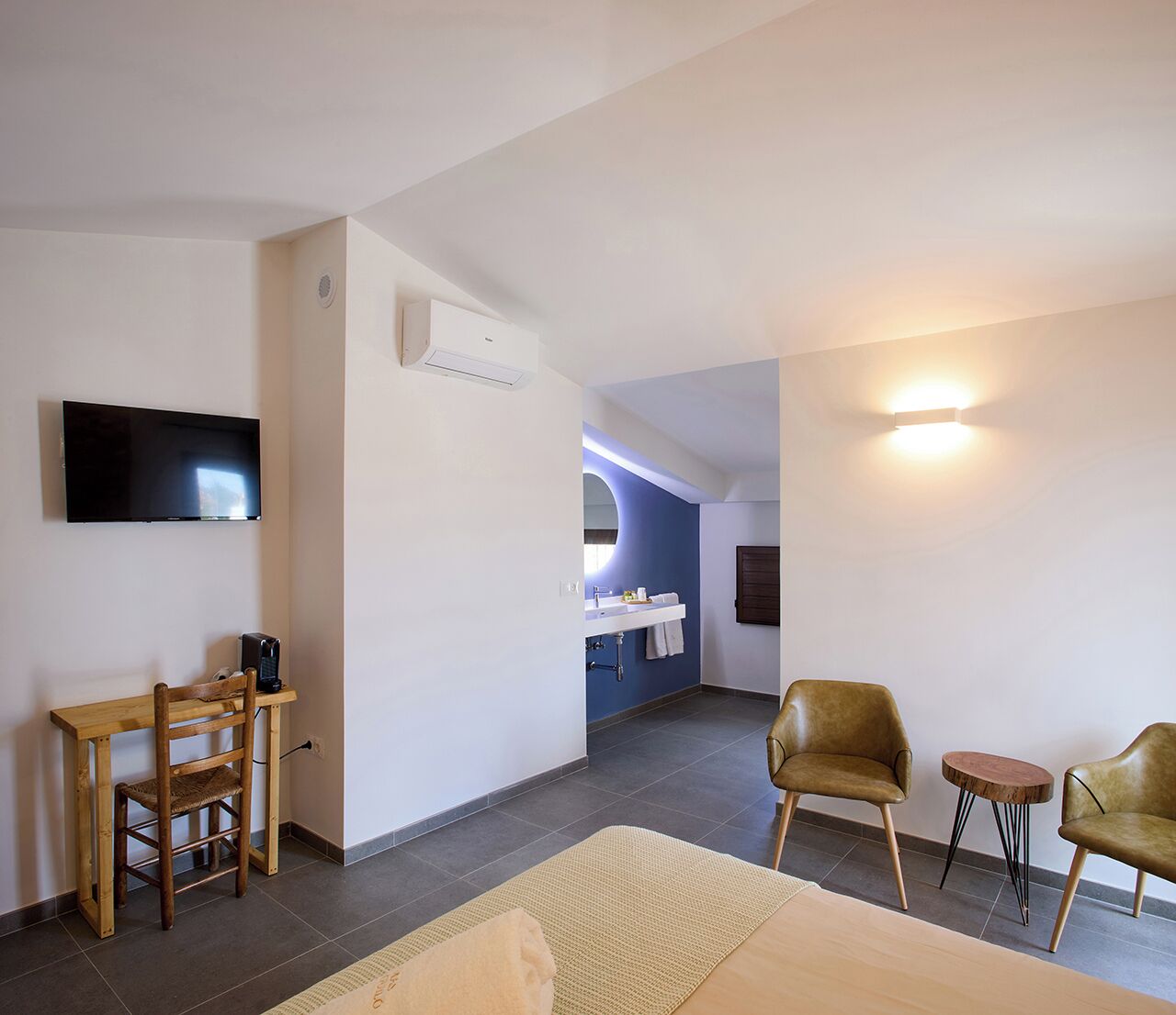 A modern hotel room with a flat-screen TV, a wooden desk, a chair, a sofa, and a bathroom visible in the background.