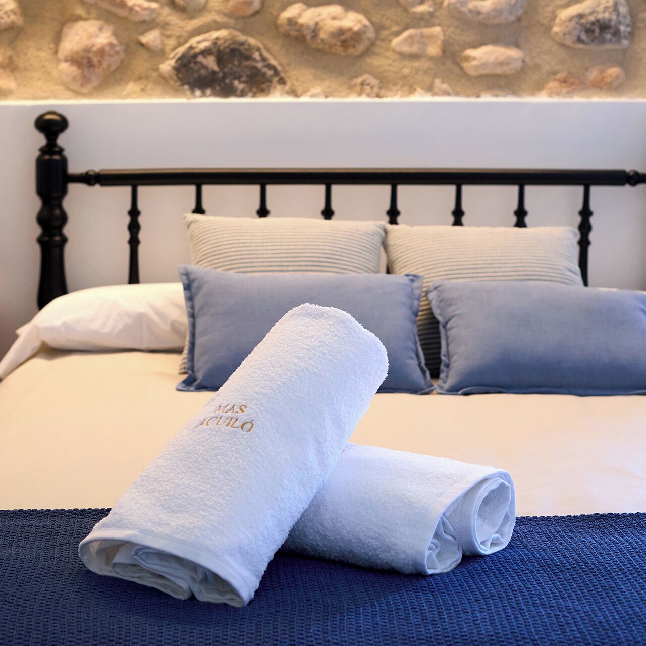 Una cama bien hecha con una toalla blanca enrollada y un tapete de baño azul, sobre un fondo de pared de piedra.