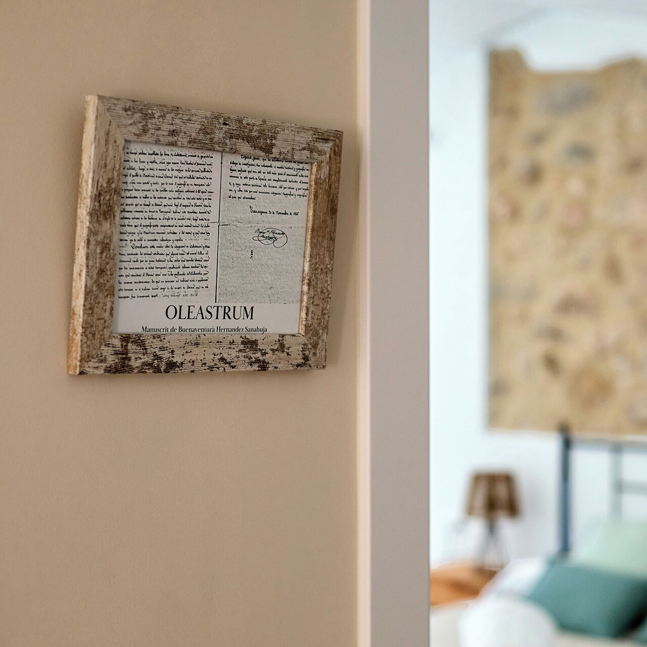Un manuscrito enmarcado con el título 'OLESTRUM' en una pared, con un fondo borroso que sugiere un dormitorio con cama y lámpara.