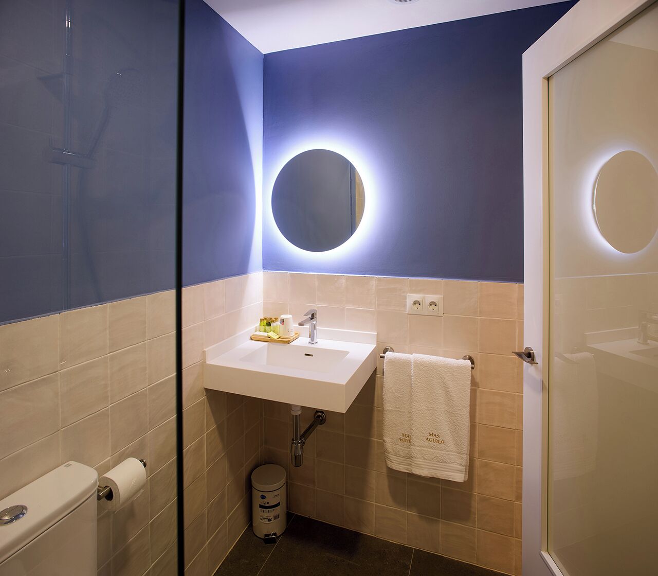 Un baño con un lavamanos blanco, un espejo con luz circular y una toalla colgada en la pared.