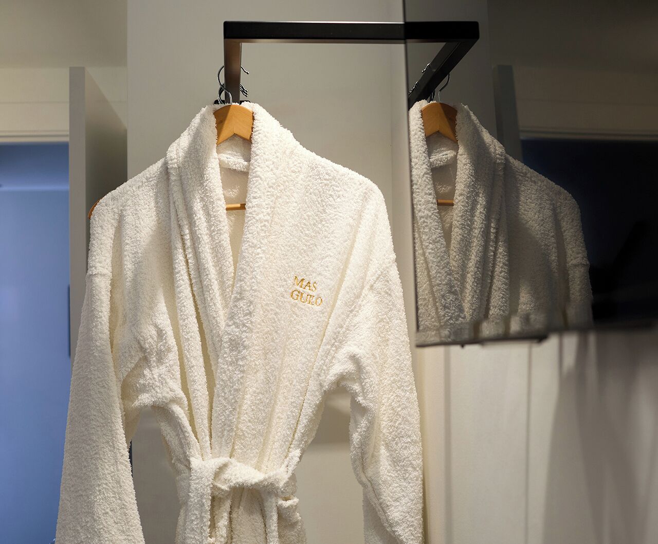A white robe with "MAS GUERO" embroidered on it, hanging on a wooden hanger.