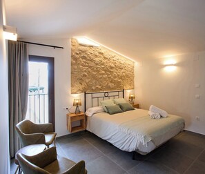 Une chambre à coucher avec un lit, deux tables de chevet avec des lampes, une chaise, un mur en pierre et une vue sur le balcon.