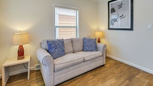 Una sala de estar con un sofá color beige, cojines estampados, una mesita con una lámpara, una ventana con persianas y un cuadro en la pared.