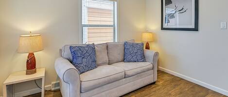 Una sala de estar con un sofá color beige, cojines estampados, una mesita con una lámpara, una ventana con persianas y un cuadro en la pared.
