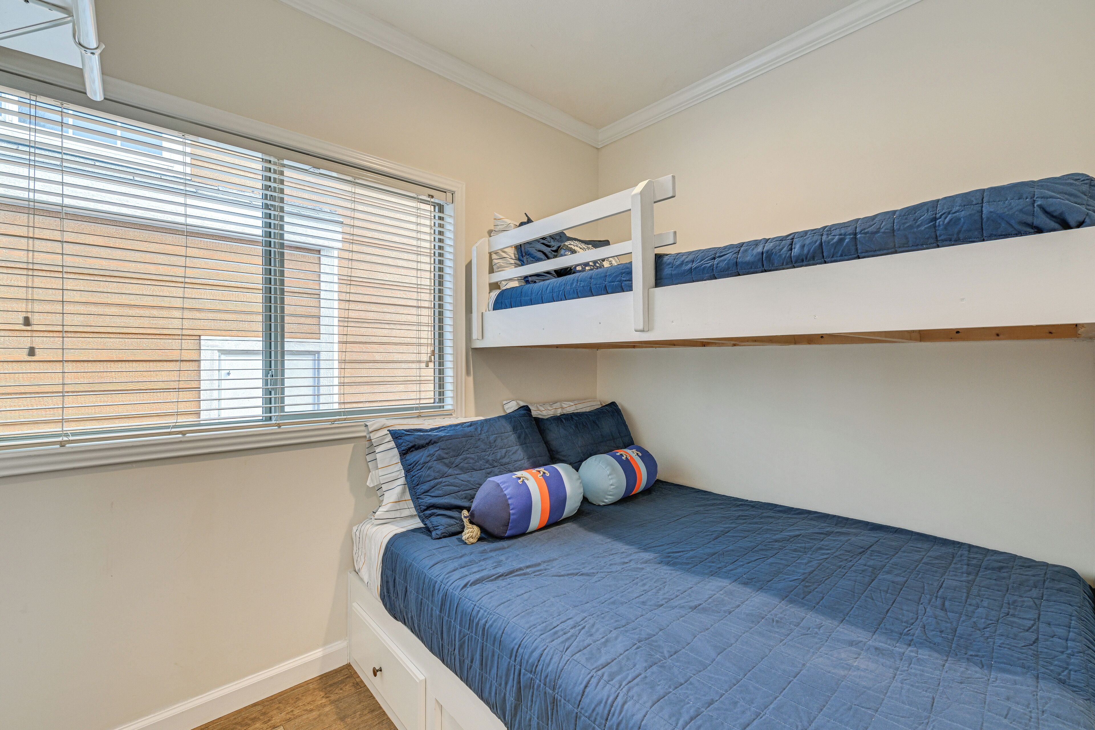 A bunk bed with a blue mattress and pillows, a window with blinds, and a white wall.