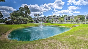 Un bassin orné d’une fontaine, entouré d’une pelouse bien entretenue et bordé d’une rangée de maisons de ville.