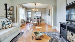 A living room with a sofa, a coffee table with a bowl, a TV, and a dining area with chairs and a table. - Walk to Beach + Lake Nokomis: Minneapolis Home! (Minneapolis)