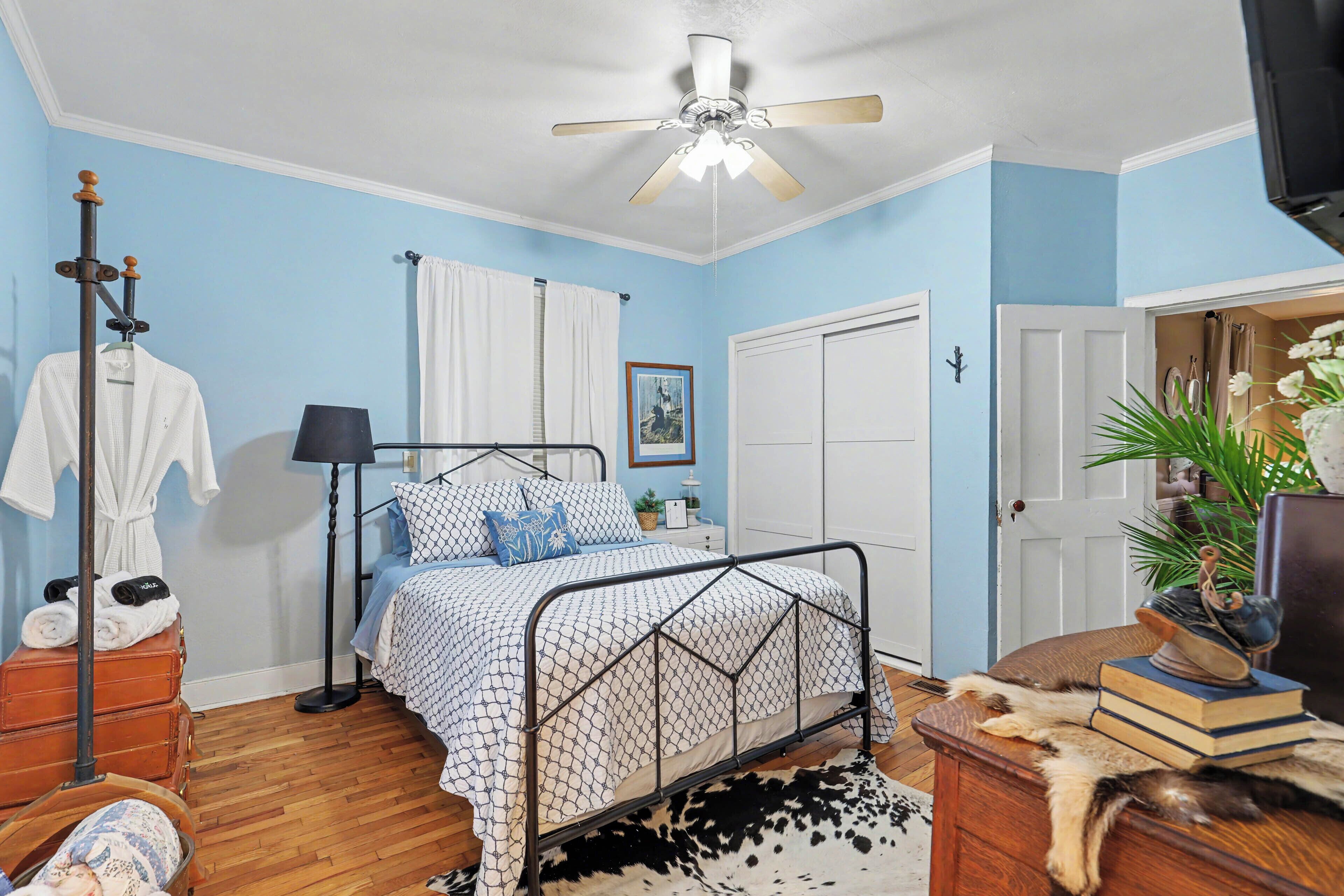 A bedroom with a bed, a ceiling fan, a wardrobe, a bedside table, a lamp, a plant, and a suitcase.