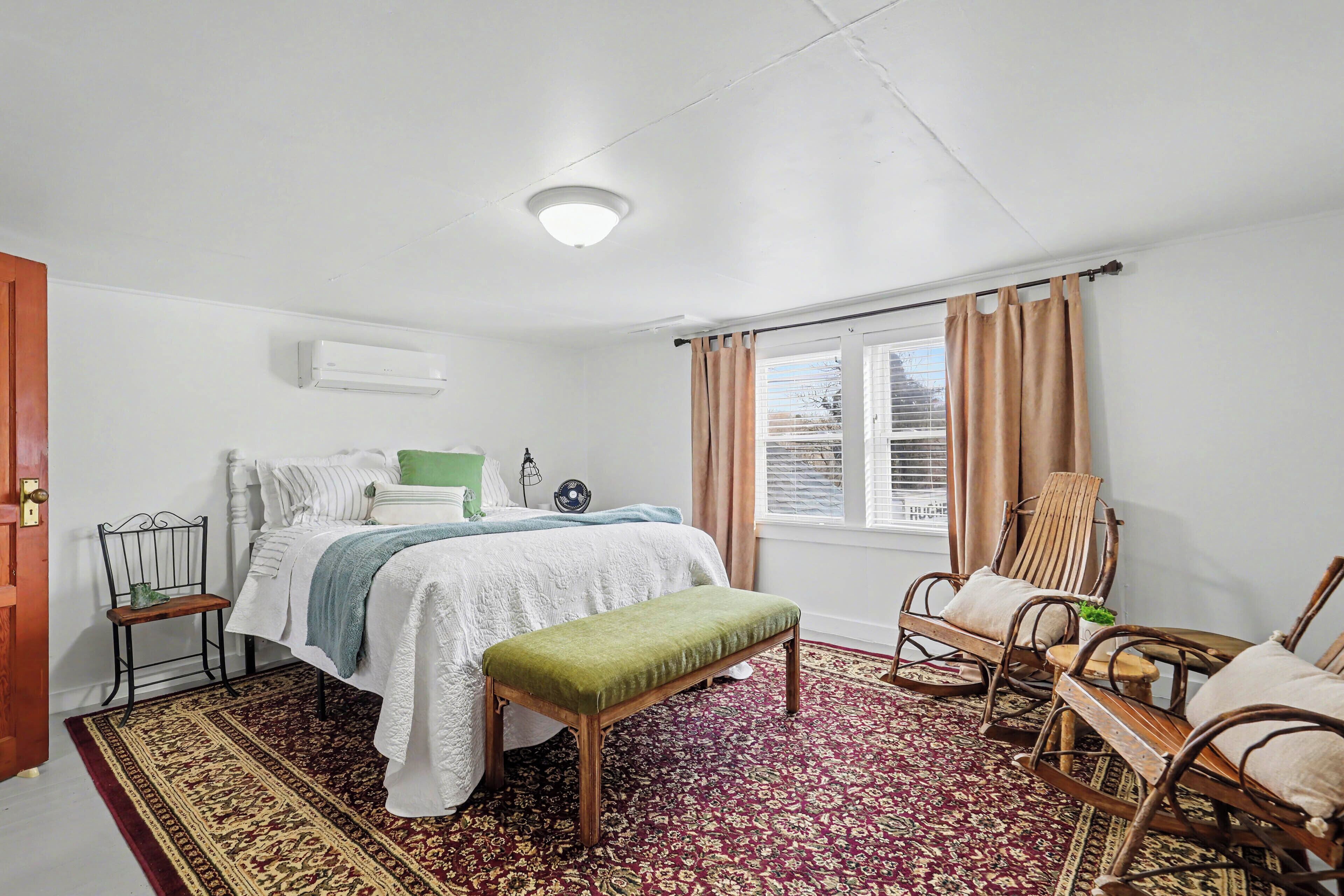 Un dormitorio con cama, un banco, sillas mecedoras, un ventilador y una ventana con cortinas.