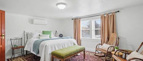 Un dormitorio con cama, un banco, sillas mecedoras, un ventilador y una ventana con cortinas.