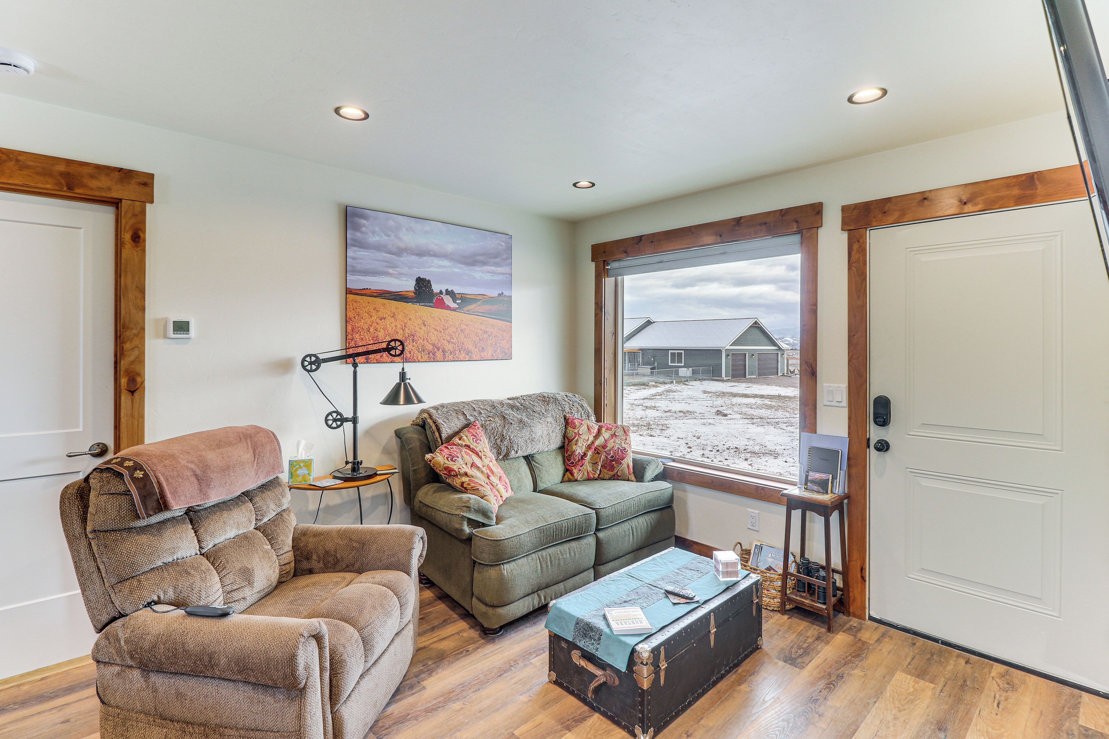 A cozy living room with a sofa, armchair, and a coffee table. There is a painting on the wall and a window with a view of a house.