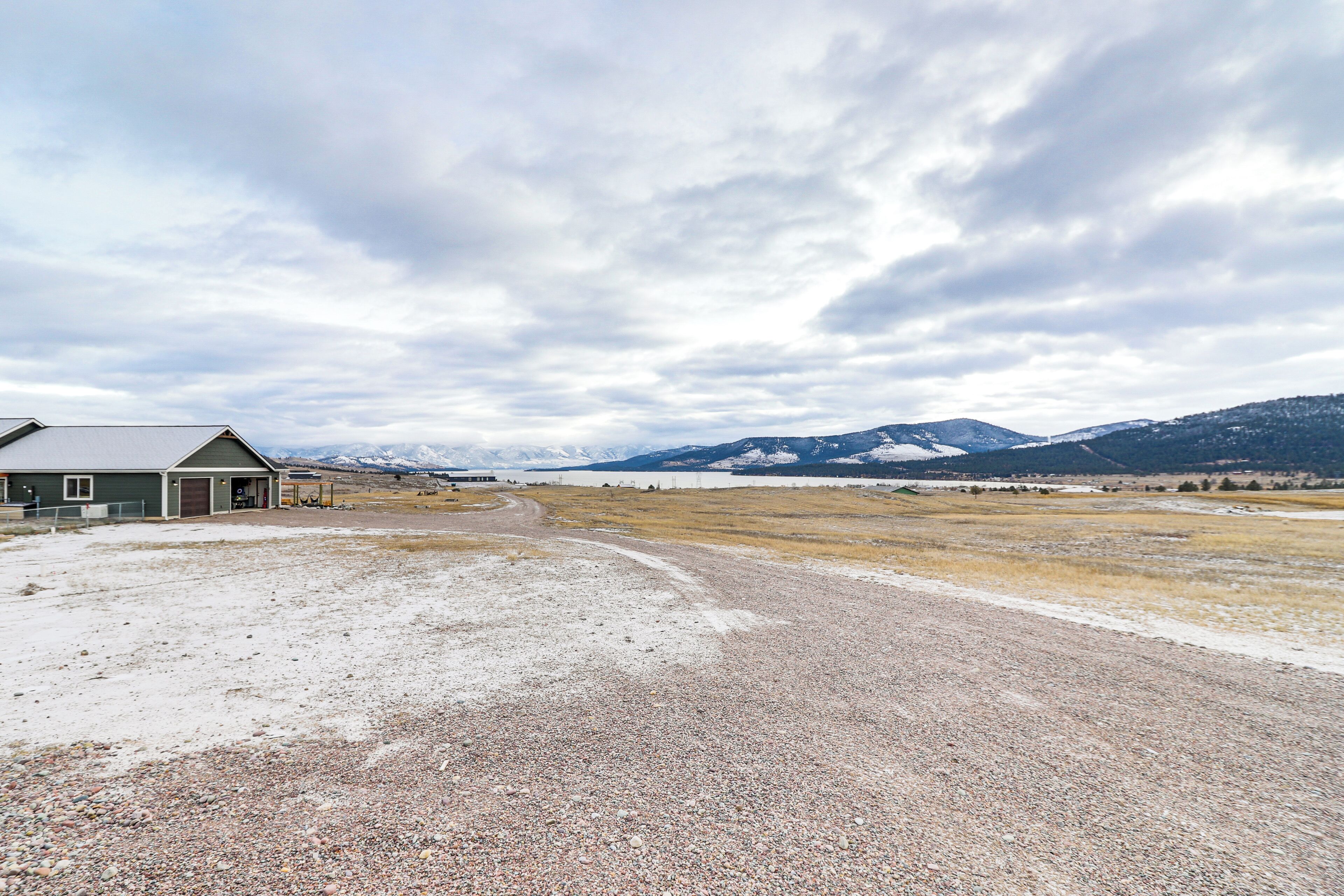 A gravel road leading to a building with a green roof, surrounded by a vast open field and distant mountains under a cloudy sky.