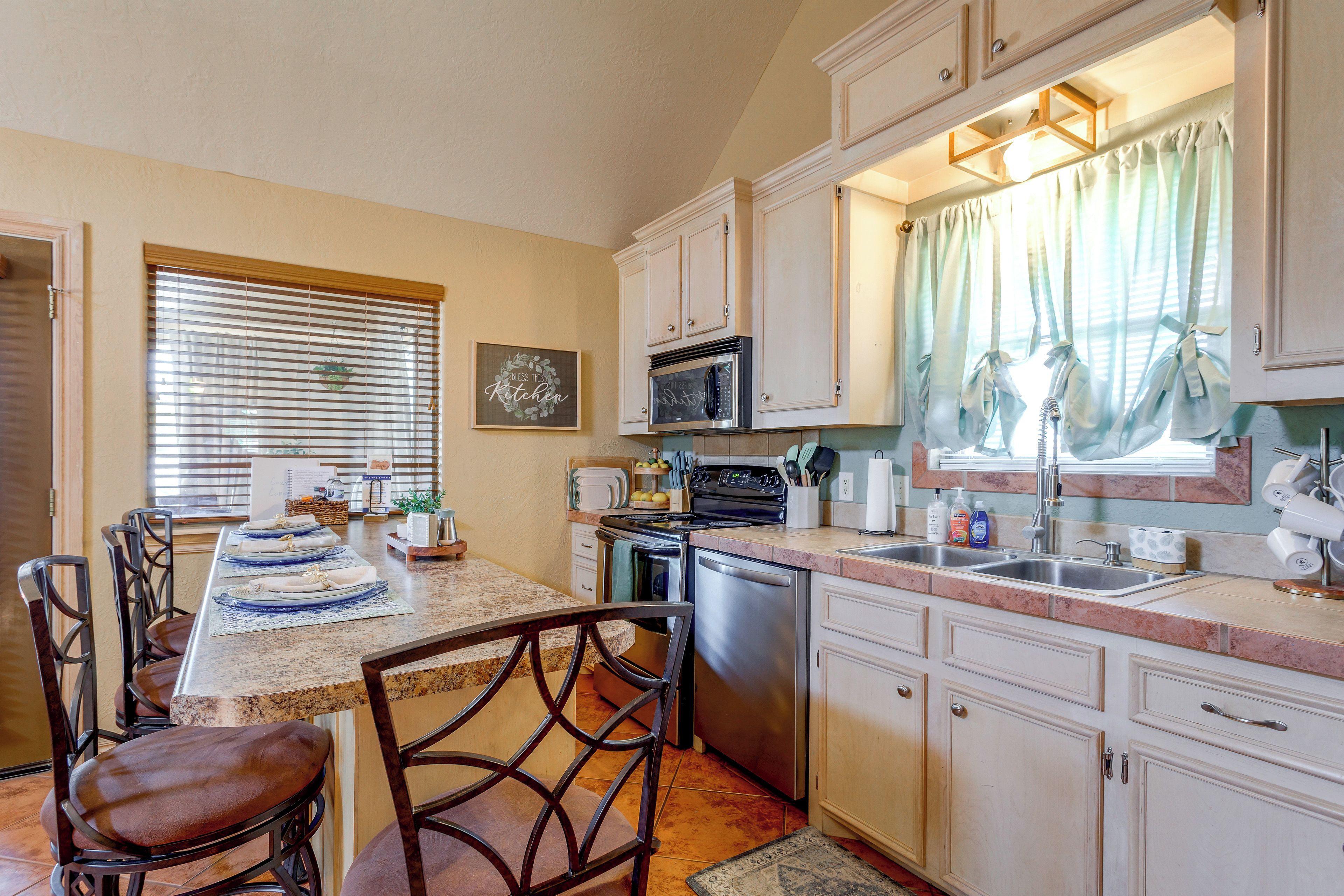 A kitchen with white cabinets, a stove, microwave, and a dishwasher. There is a dining table with chairs and a window with blinds.