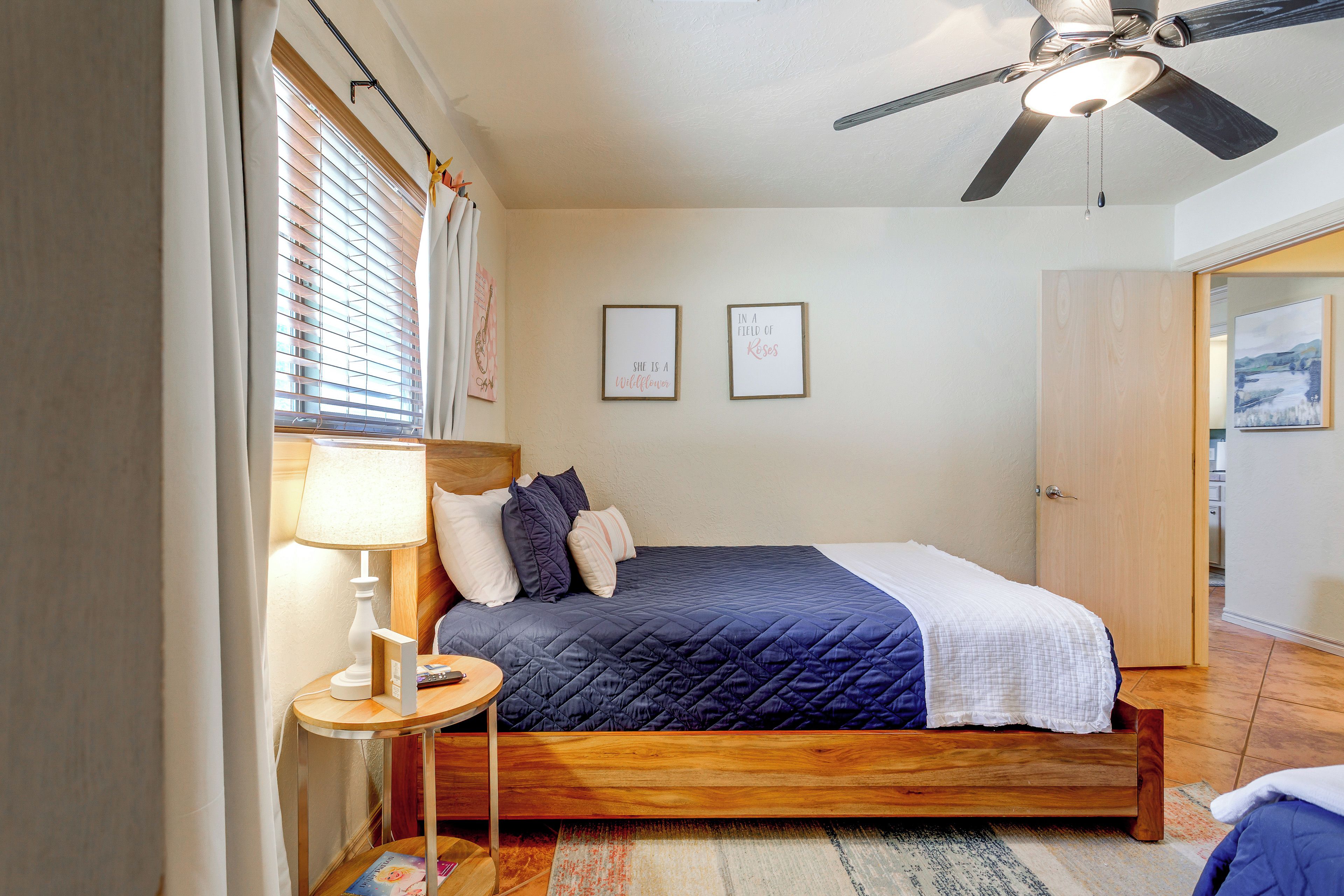 A bedroom with a bed, a bedside table with a lamp, a ceiling fan, and two framed pictures on the wall.