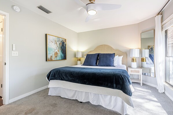 A bedroom with a bed, bedside lamps, a ceiling fan, a painting, and a window with curtains.