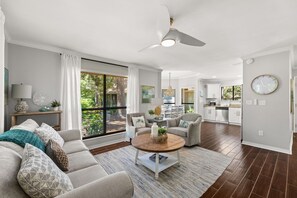 A spacious living room with a large window, a ceiling fan, and a kitchen in the background.