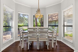 A dining area with a round table and six chairs, surrounded by large windows with blinds.