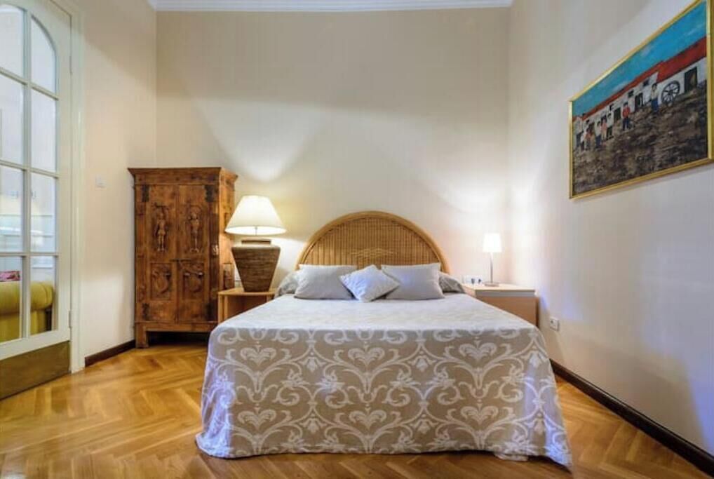 A bedroom with a wooden floor, a bed with a patterned bedspread, a wooden wardrobe, and a painting on the wall.