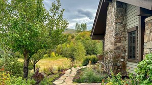 A stone pathway leading to a stone building with a chimney, surrounded by lush greenery and a hilly landscape.