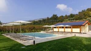 A swimming pool with lounge chairs and umbrellas, a building with a solar panel roof, and a grassy area with trees in the background. - Agriturismo Agricamping GARDA NATURA (Costermano sul Garda)