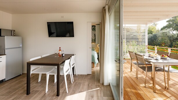 A modern kitchen with a flat-screen TV, a dining area with a table and chairs, and an outdoor deck with a wooden floor. - Agriturismo Agricamping GARDA NATURA (Costermano sul Garda)