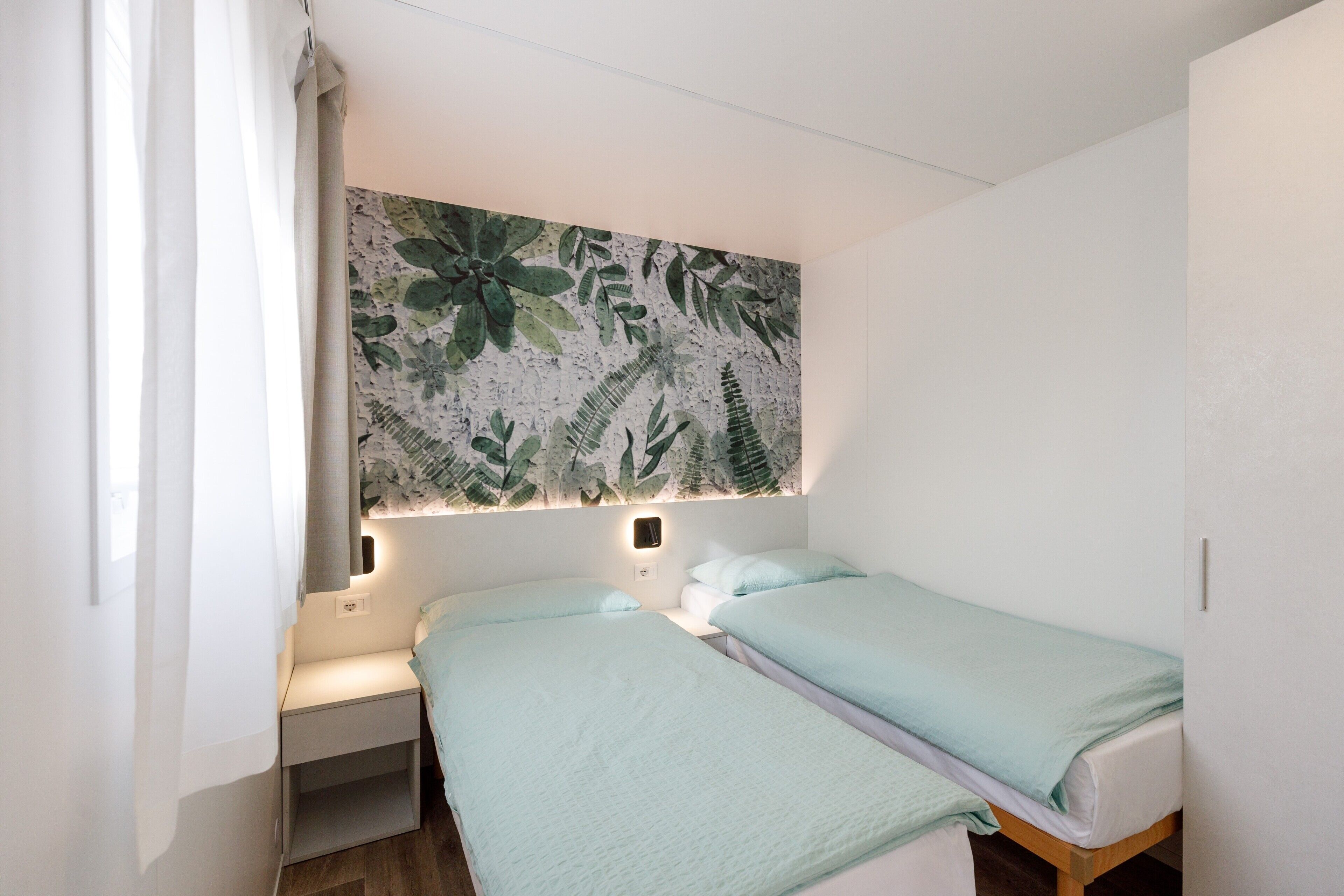 A bedroom with two beds, a nightstand, a wall-mounted light, and a large tropical leaf-patterned wall hanging.