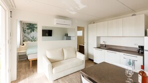 A modern kitchen with white cabinets, a built-in oven, and a coffee maker. There is a beige sofa in the living area and a dining table with a wine glass and bottle. - Agriturismo Agricamping GARDA NATURA (Costermano sul Garda)