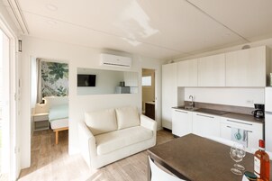 Una cucina moderna con mobili bianchi, forno integrato e macchina per il caffè. Nell'area living, un divano color beige e un tavolo da pranzo con un bicchiere e una bottiglia di vino.