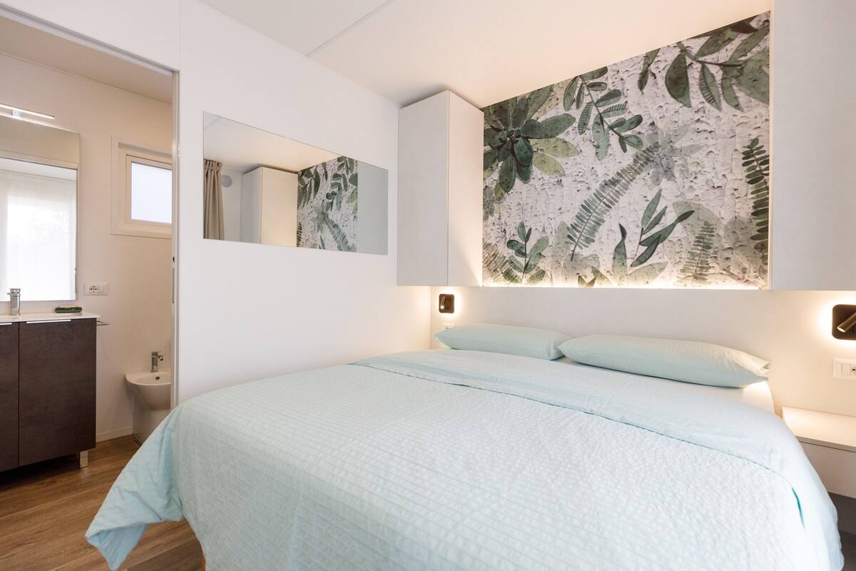 A bedroom with a bed, a bedside table, a nightstand, a mirror, and a wall-mounted artwork.