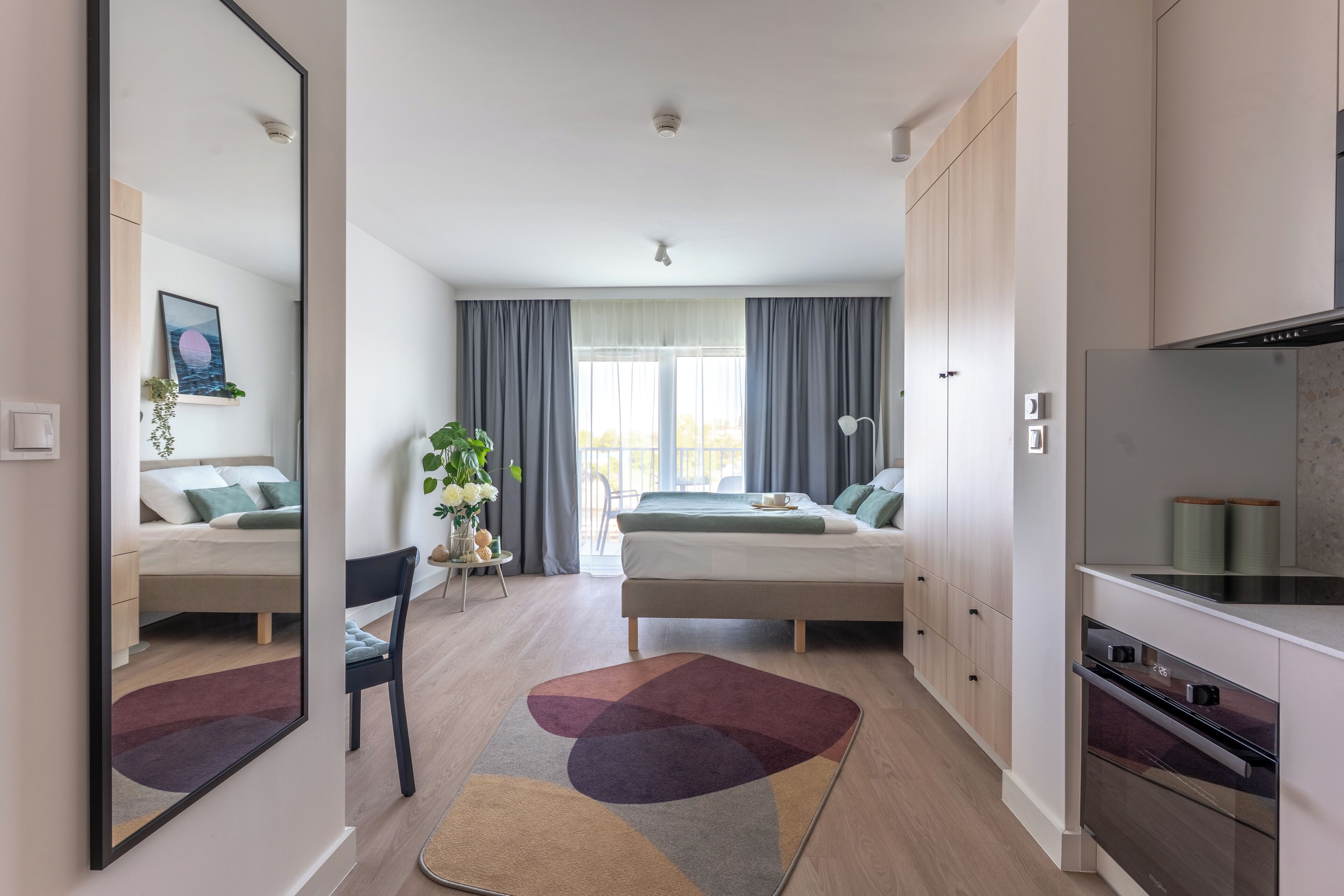 A modern hotel room with a bed, a desk, a chair, a mirror, a window with curtains, and a kitchenette with a stove and oven.