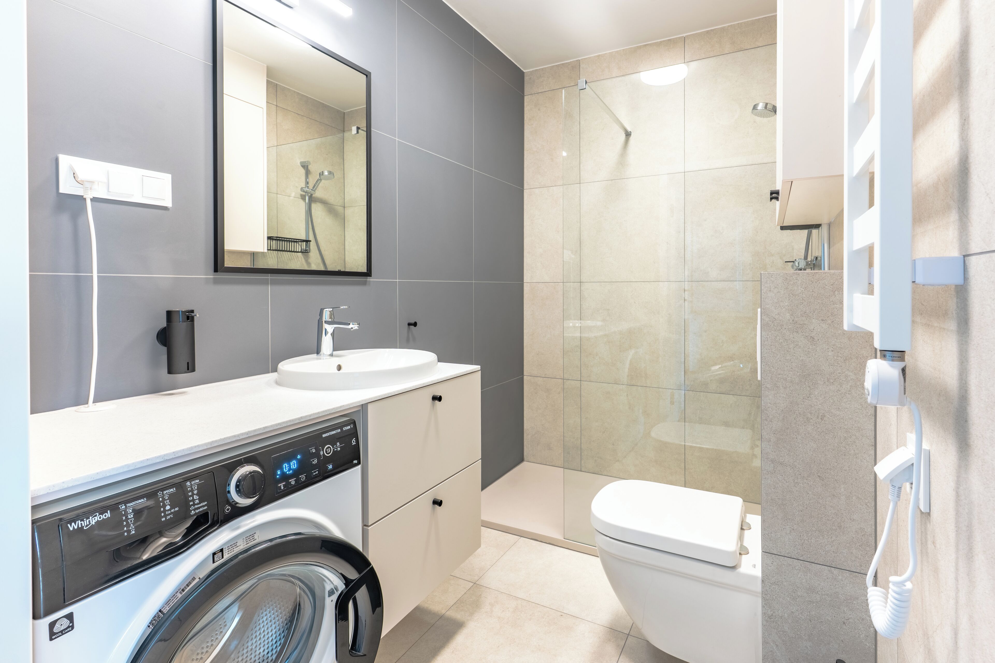 A modern bathroom with a built-in washer and dryer, a large mirror, a toilet, and a glass shower enclosure.