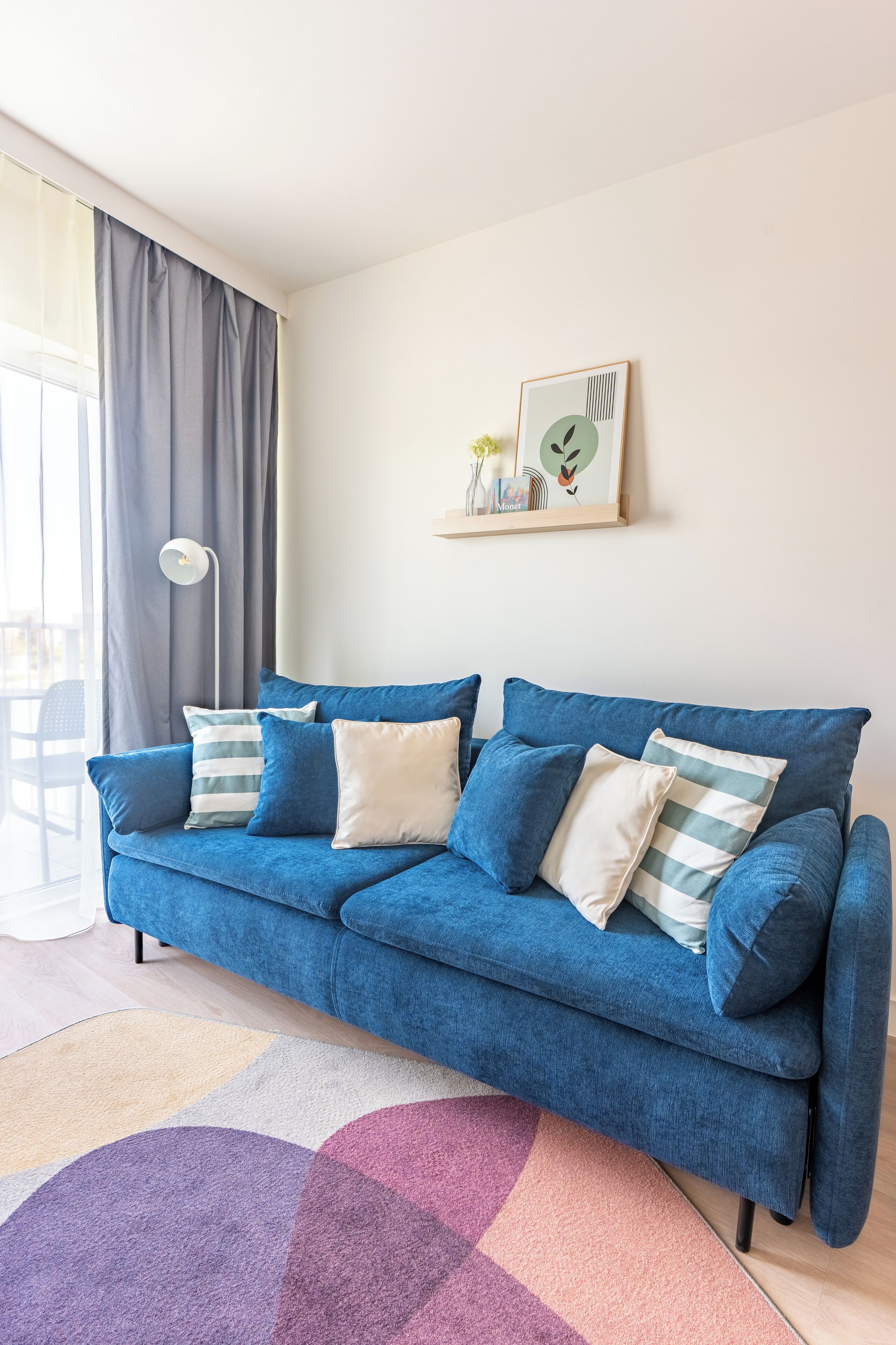 A blue sofa with striped pillows, a colorful rug, a framed picture on the wall, and a window with curtains.