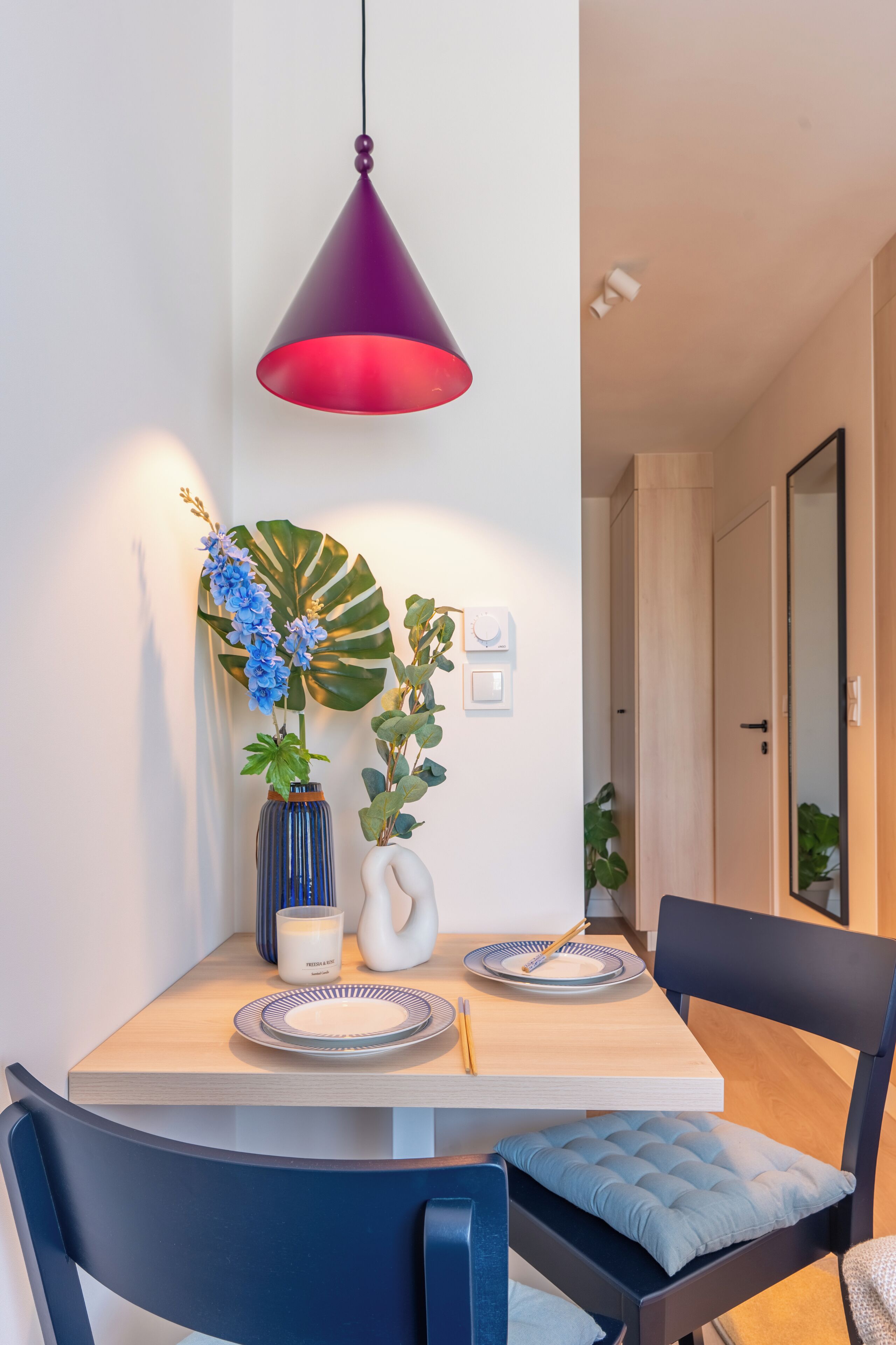 A dining area with a wooden table, two chairs, a vase with blue flowers, a potted plant, and a pendant light.