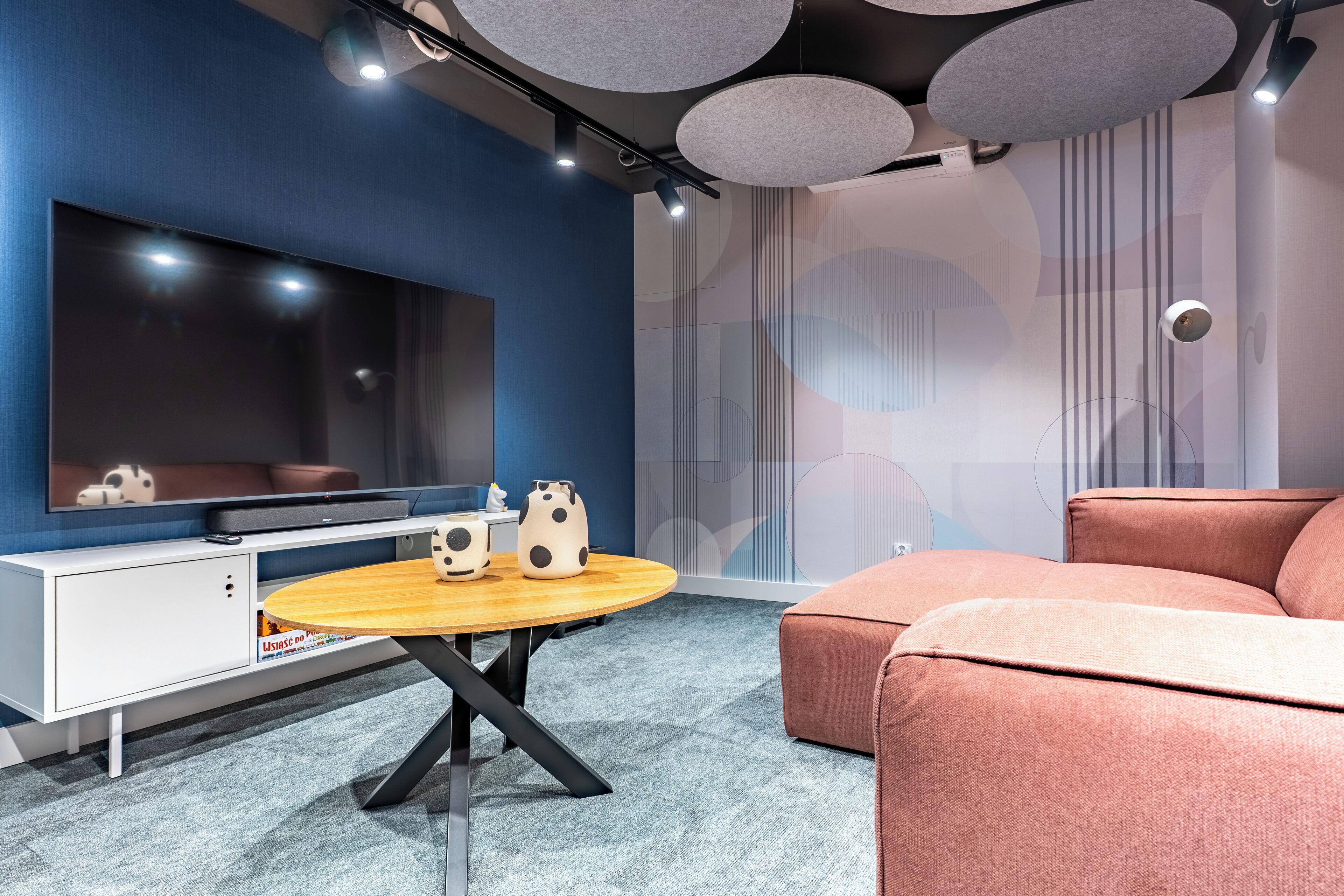 Una sala de estar moderna con un televisor de pantalla plana, una mesa redonda de madera, dos adornos con forma de cerdo, un sofá rosa y una pared con patrones abstractos.