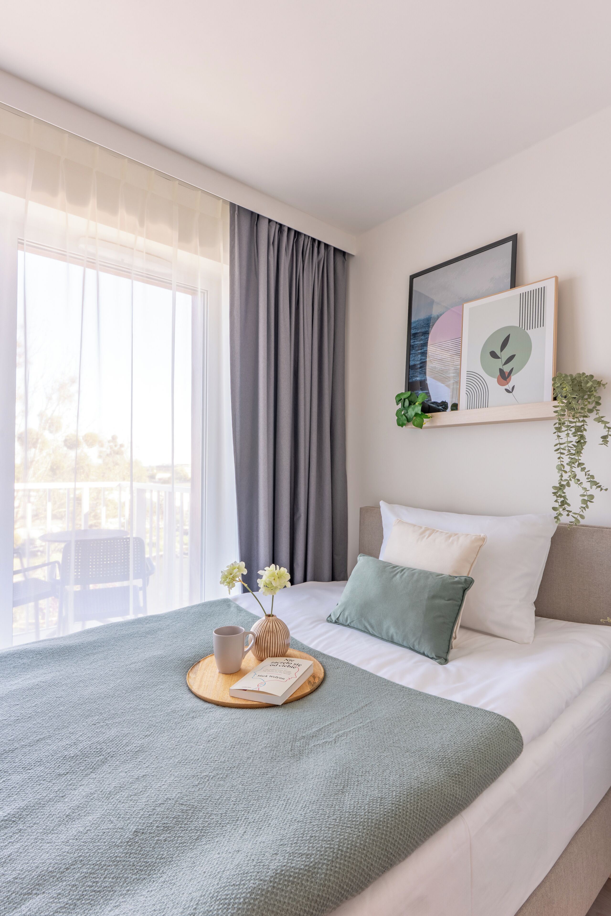 A neatly made bed with a green blanket, a white pillow, and a grey pillow. There is a framed picture on the wall, a small plant, and a window with grey curtains.
